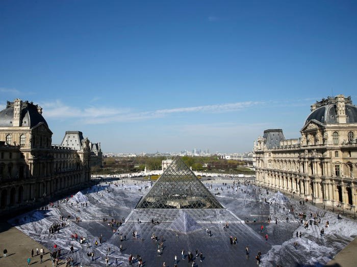 Туристы гуляют по гигантским фотографиям в главном дворе Лувра.